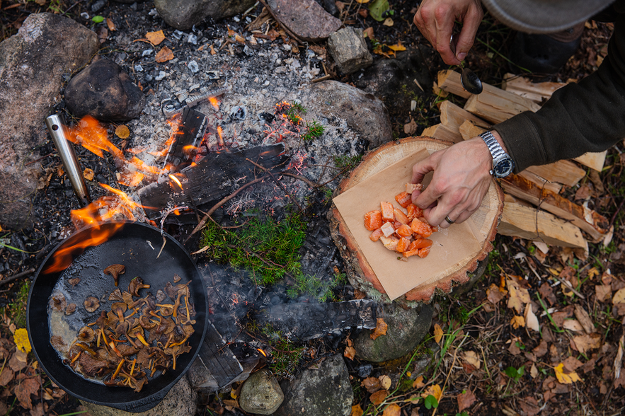 Eine Pfanne mit Pilzen, die über offenem Feuer auf dem Waldboden gebraten werden. Und ein Baumstumpf, der als Schneidebrett dient.