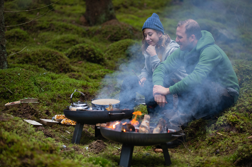 Ein Mann und eine Frau sitzen im Wald und kochen über offenem Feuer. Rauch steigt auf.